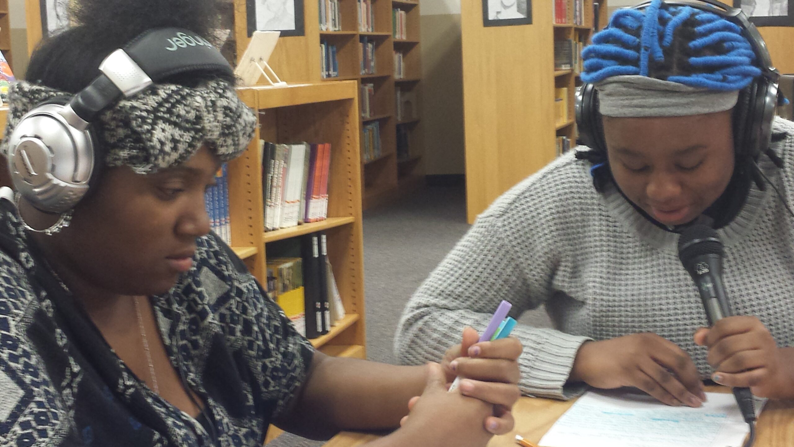 Two teens wearing headphones looking down at a piece of paper on a wooden table in a library, one speaking into a microphone.