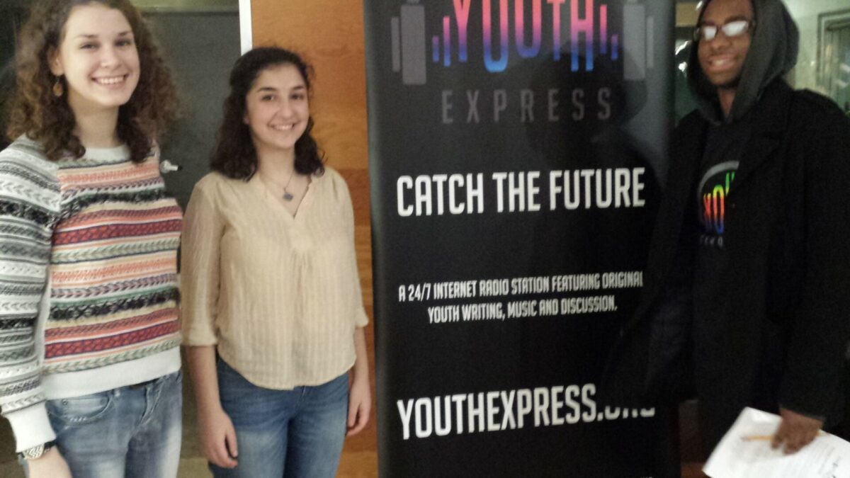 Three teens smiling and standing in next to a banner advertising Youth Express