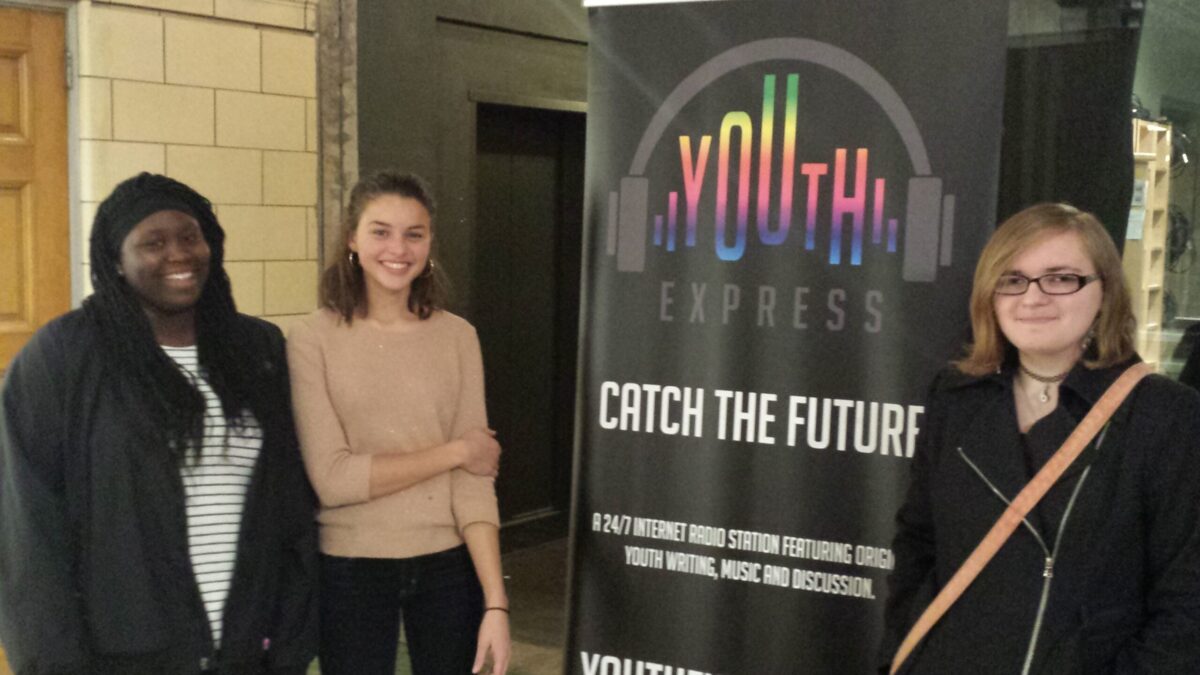 Three teens smiling and standing next to a sign advertising Youth Express