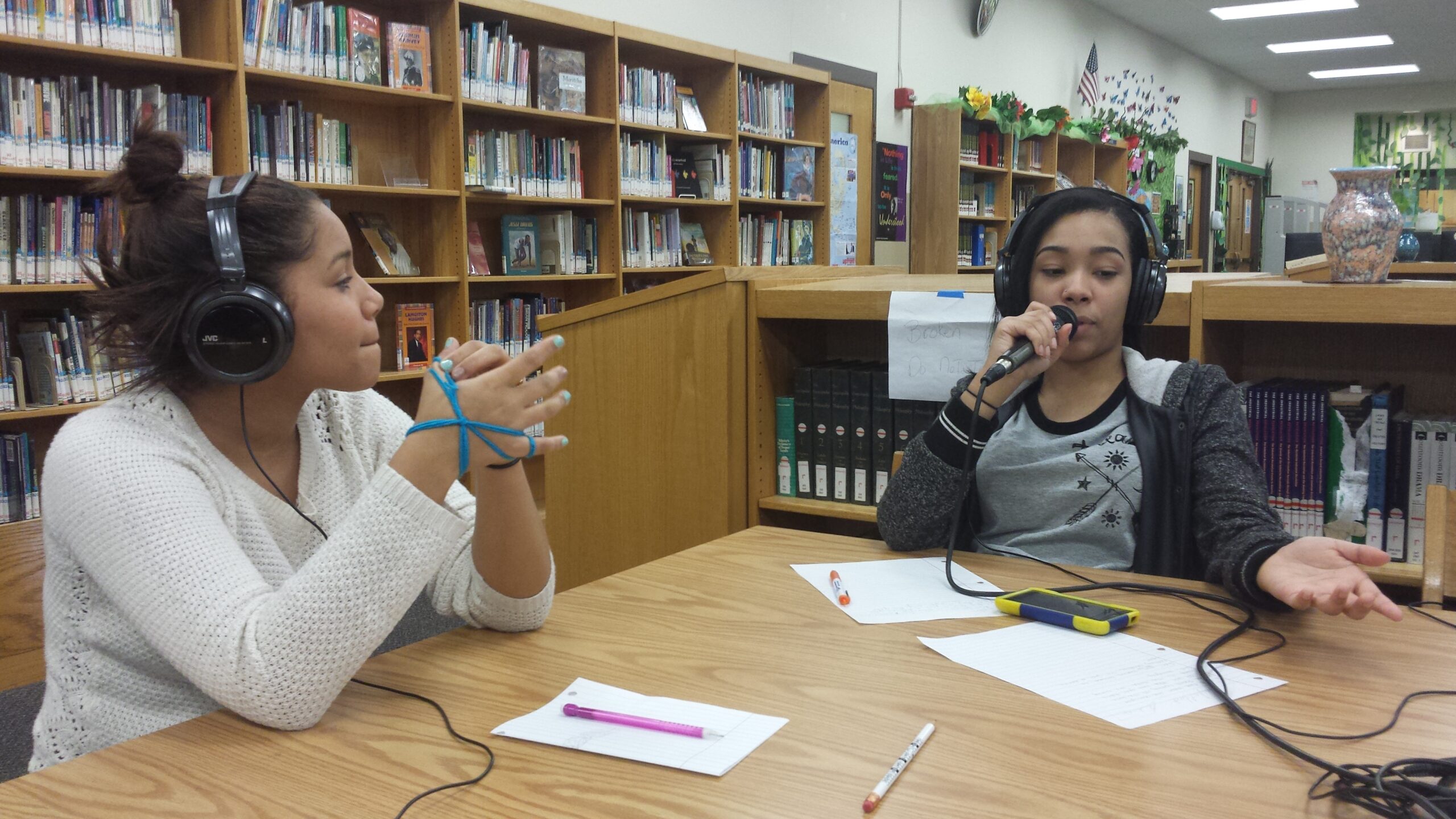 Two teens wearing headphones sitting at a wooden table in a library, one teen holding and speaking into a microphone