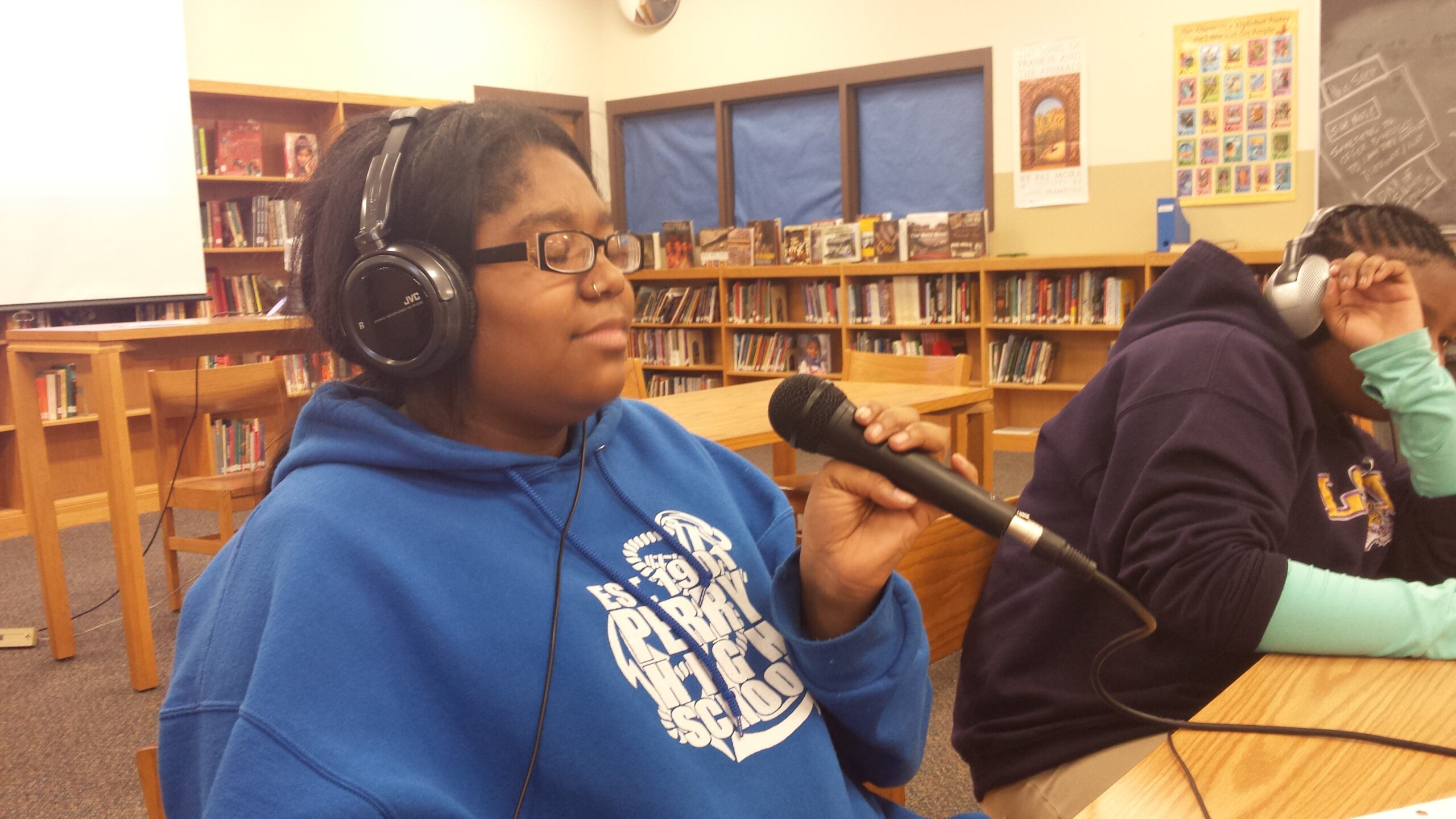 Two teens wearing headphones sitting at a wooden table in a library, one teen holding a microphone
