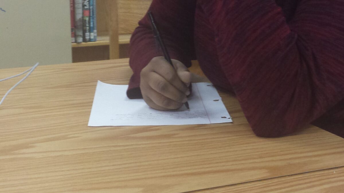 An individual writing on a piece of notebook paper on a brown table in a library