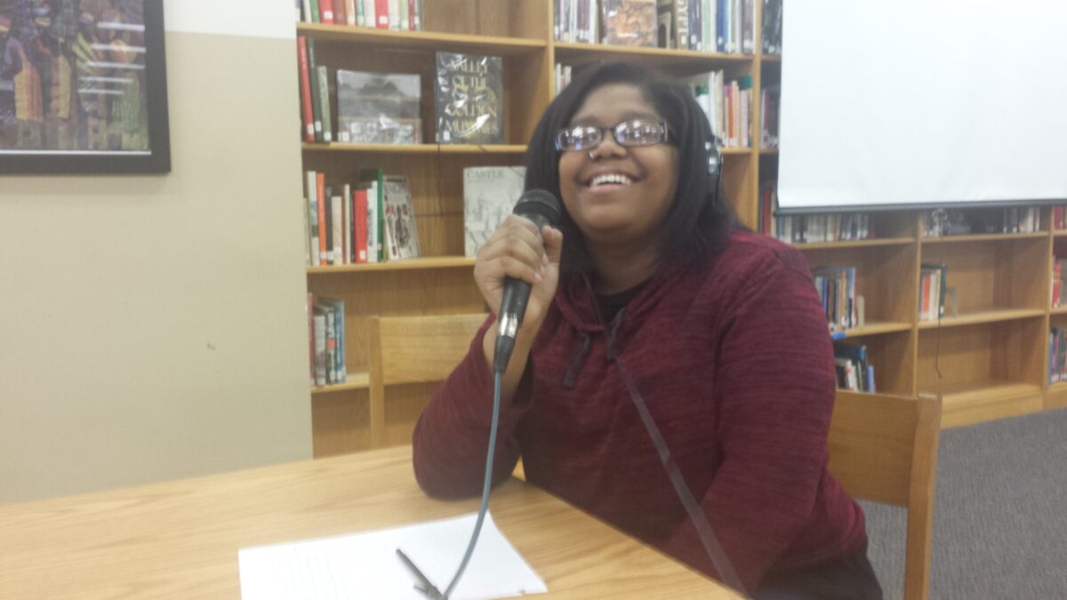 A teen wearing headphones smiling and holding a microphone sitting behind a piece of paper on a brown table in a library