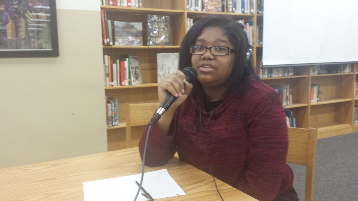 A teen wearing headphones holding and speaking into a microphone sitting behind a piece of paper on a brown table in a library