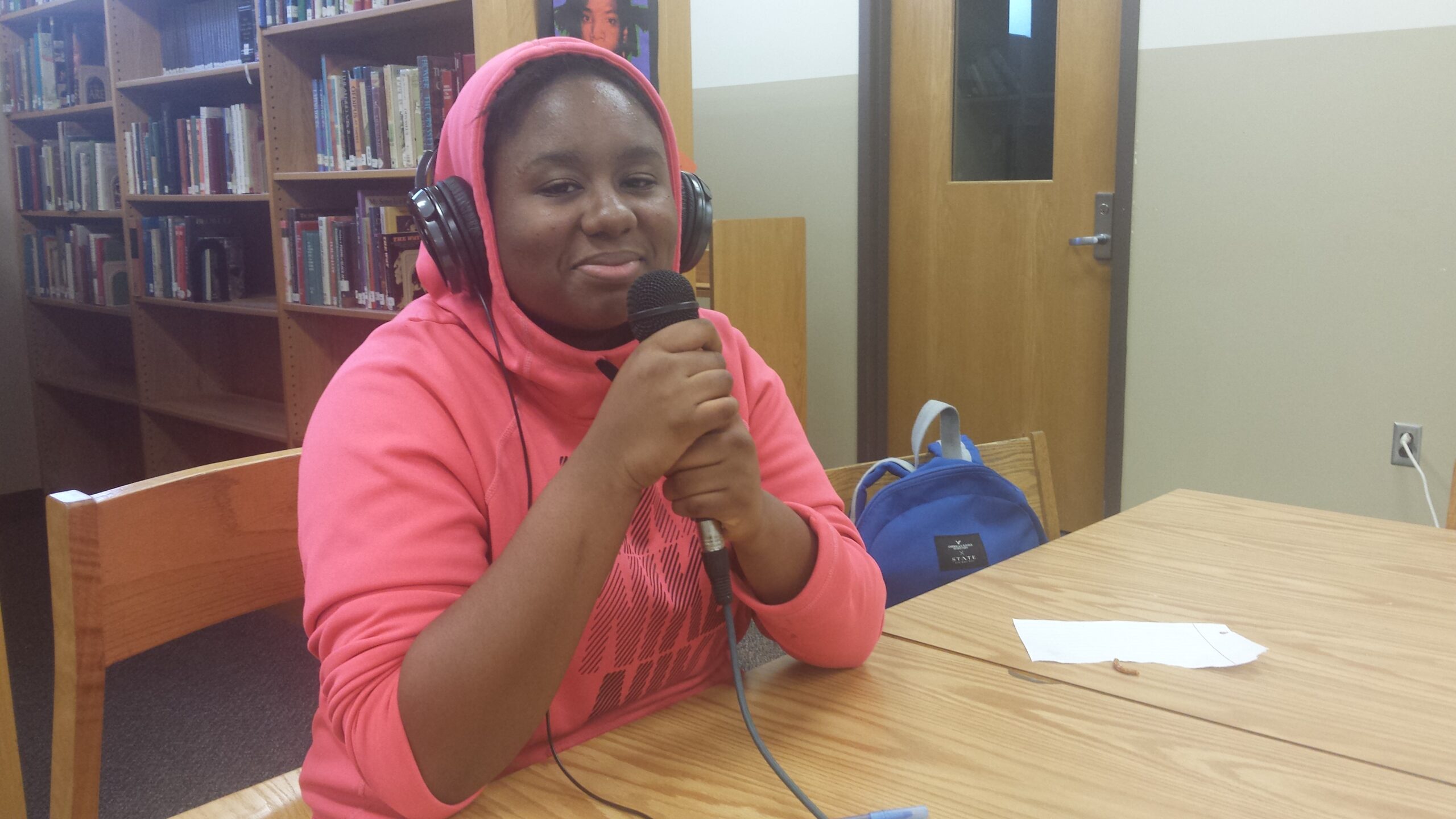 A teen wearing headphones holding a microphone sitting behind a piece of paper on a brown table in a library