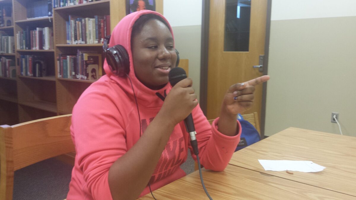 A teen wearing headphones holding and speaking into a microphone sitting behind a piece of paper on a brown table in a library