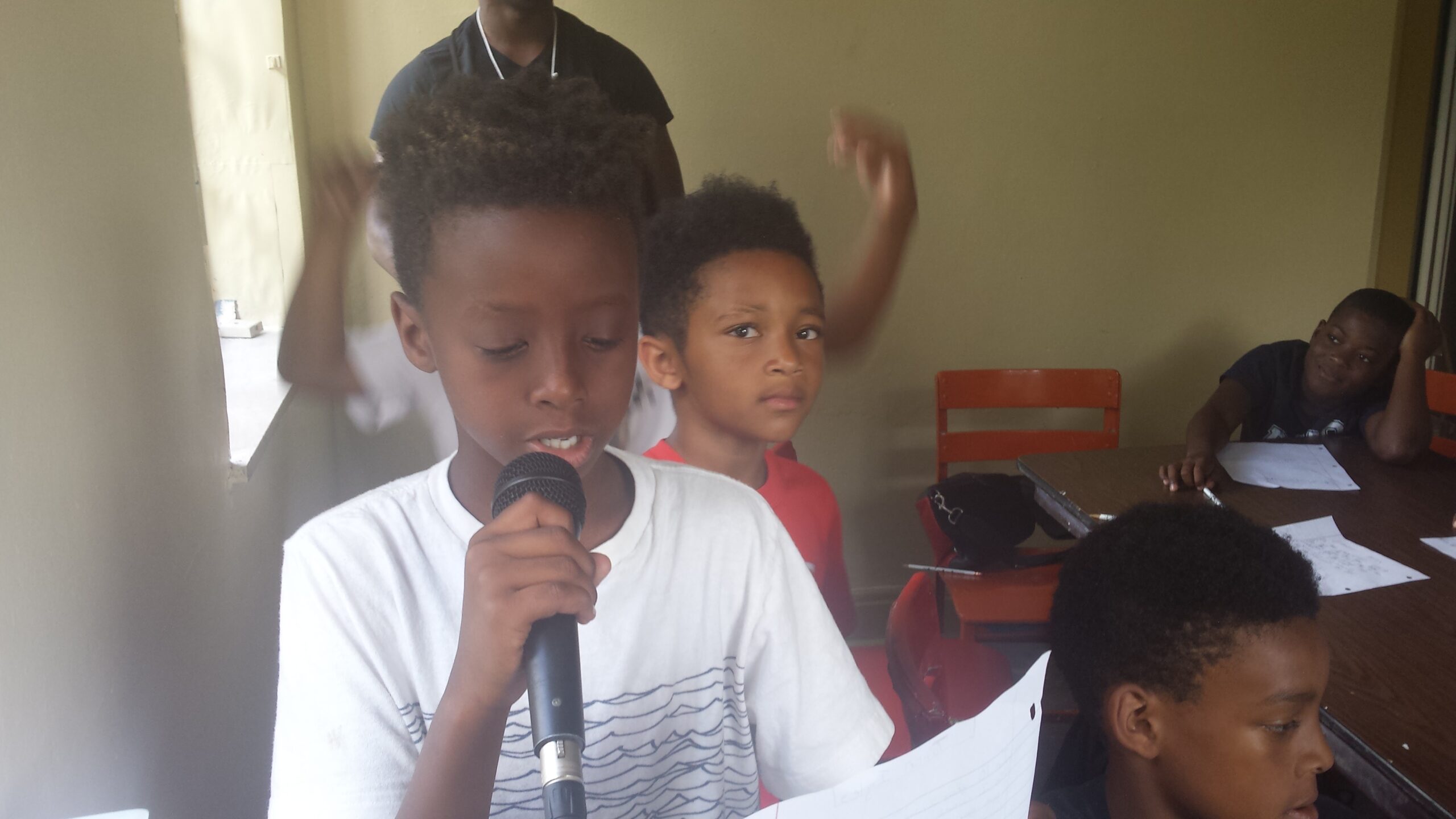 A youth holding a piece of paper and speaking into a microphone as three youth listen in the background