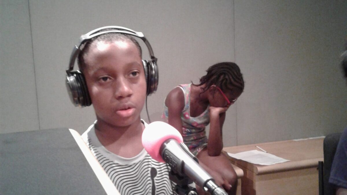 A youth wearing headphones speaking into a microphone and a youth sitting in the background in a radio studio