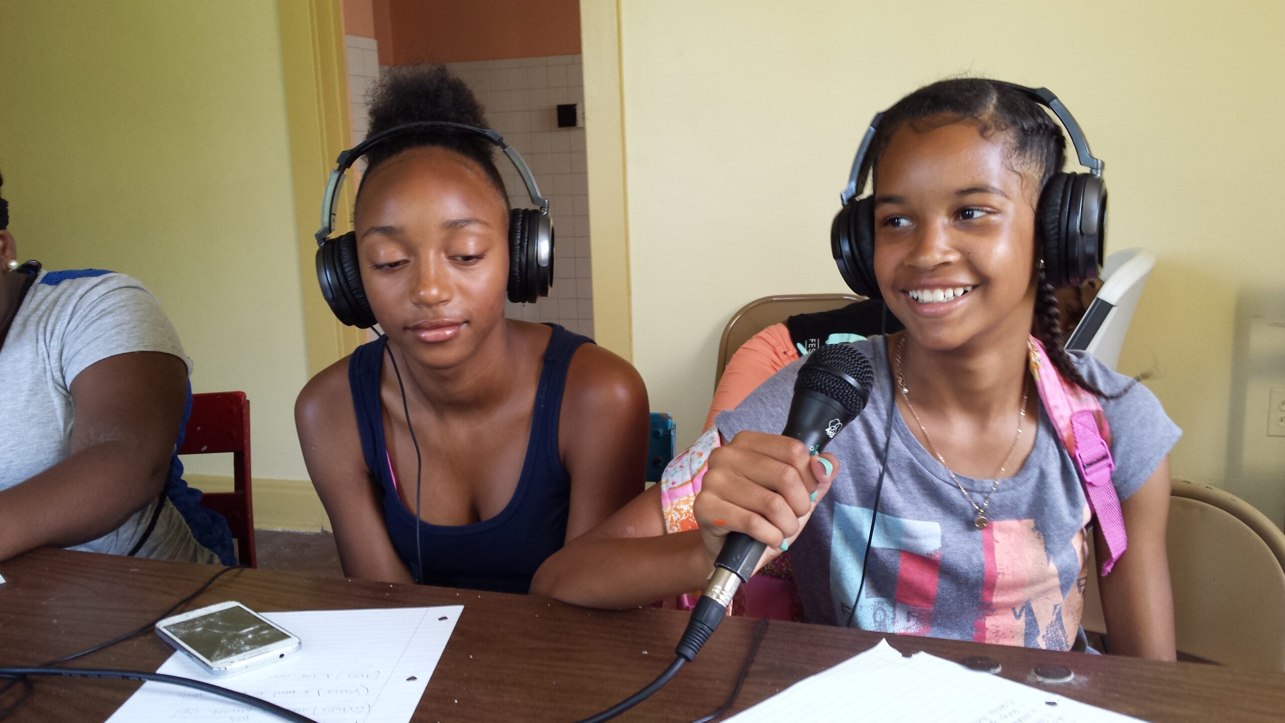 Two youth wearing headphones sitting at a brown table, one holding a microphone