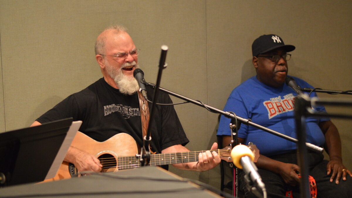 An adult playing an acoustic guitar and singing into a microphone and an adult sitting behind a microphone holding a tambourine in a radio studio