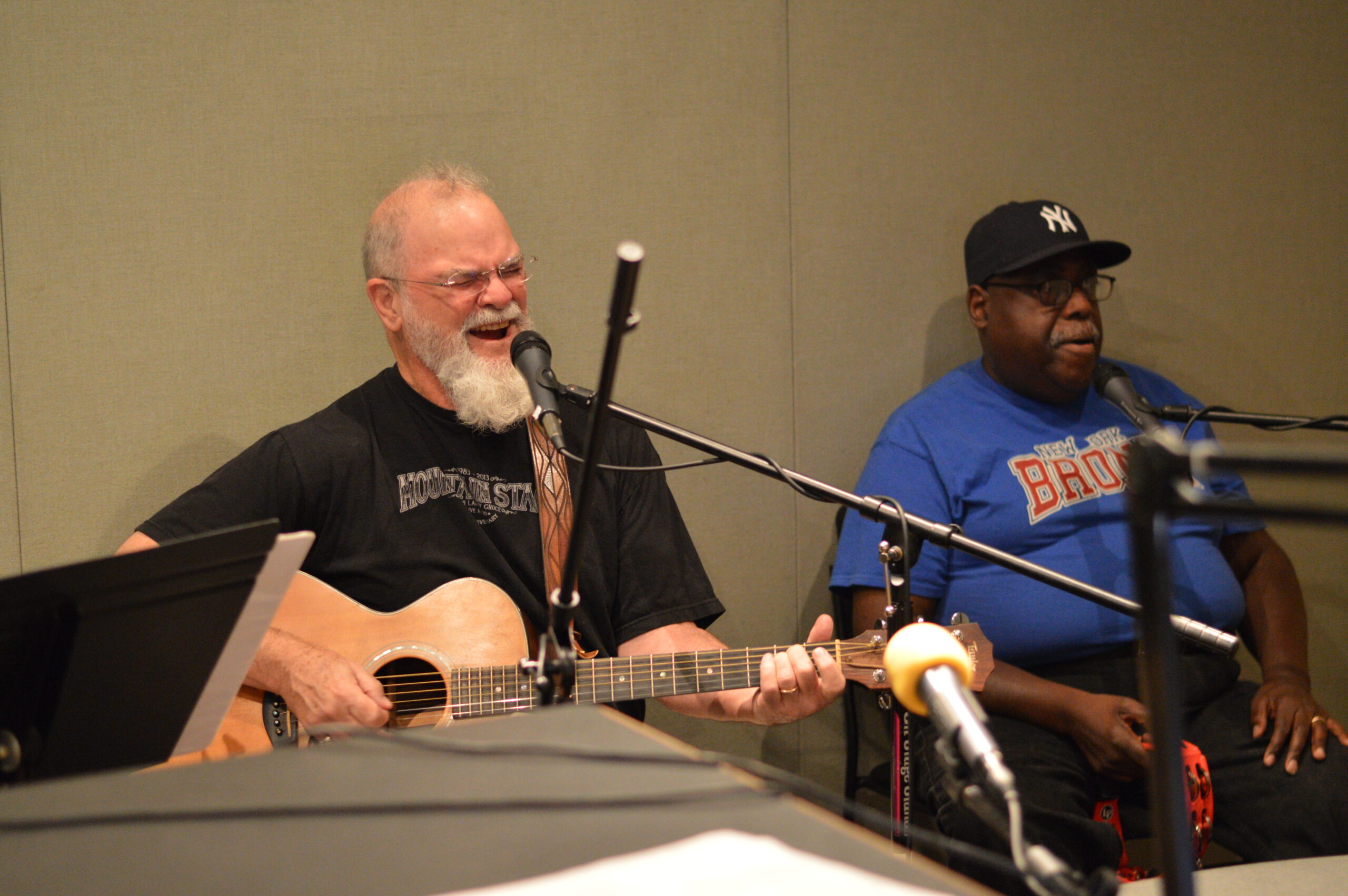 An adult playing an acoustic guitar and singing into a microphone and an adult sitting behind a microphone holding a tambourine in a radio studio