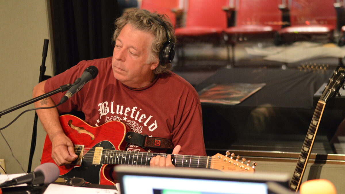 An adult wearing headphones playing an electric guitar sitting behind a microphone in a radio studio