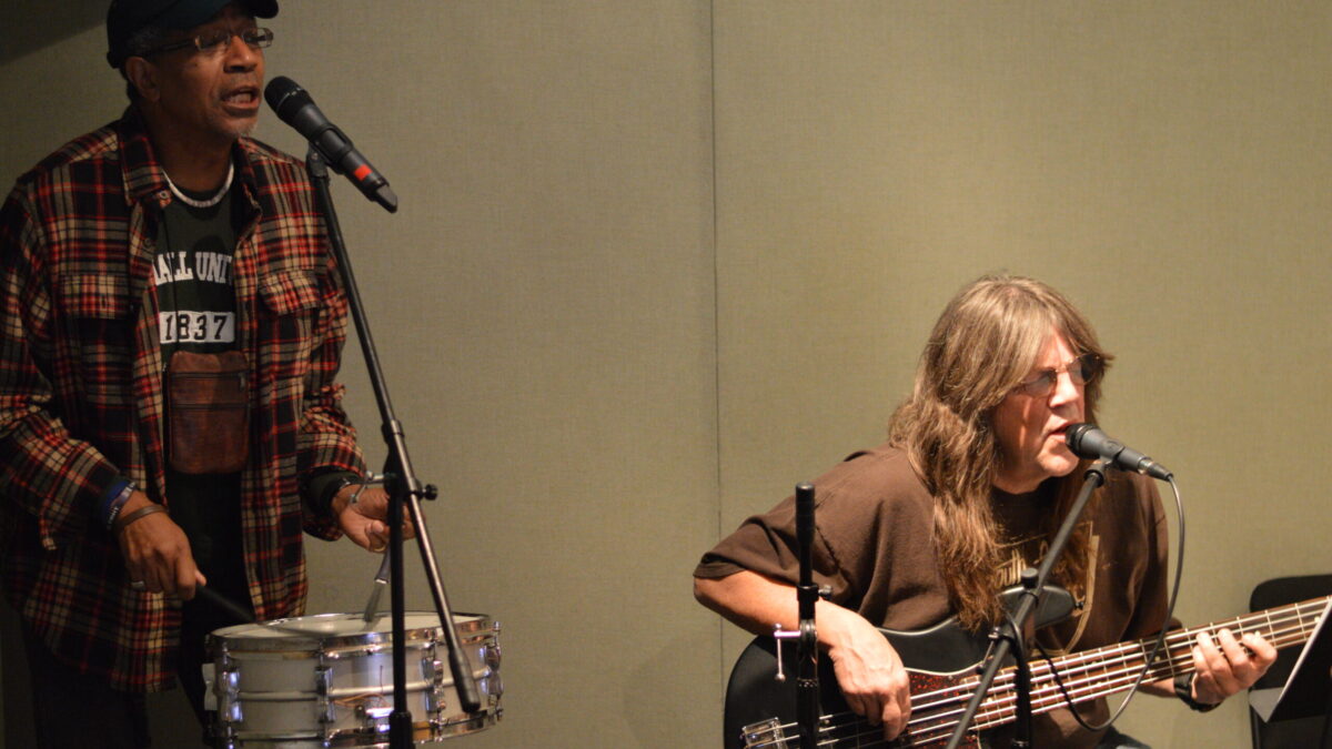 An adult playing a snare drum and an adult playing an electric bass in a radio studio