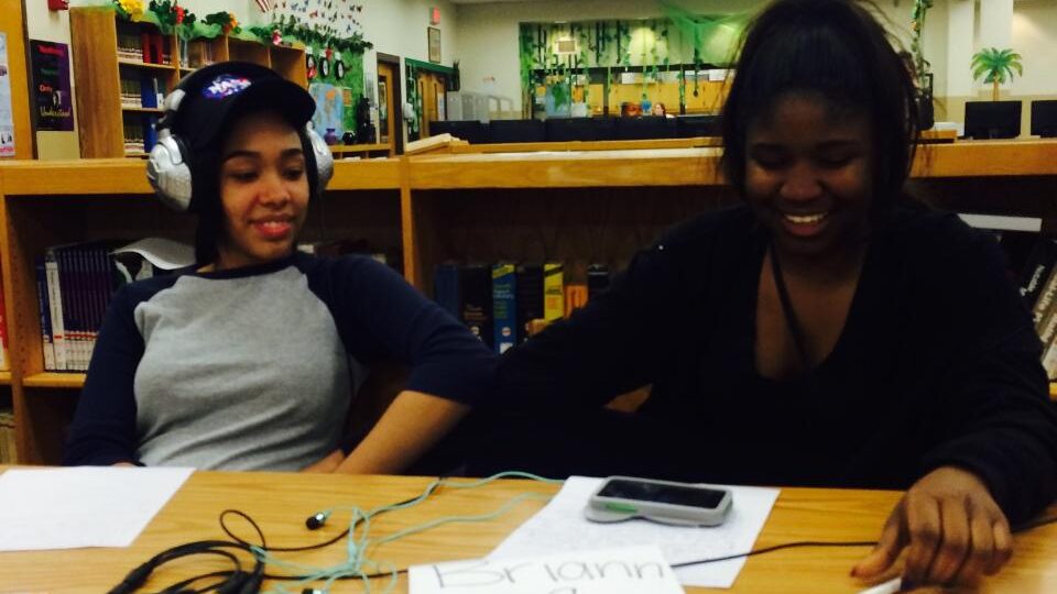 FullSizeRender (2) Two teens wearing headphones looking down at a smartphone on a piece of paper on a wooden table in a library