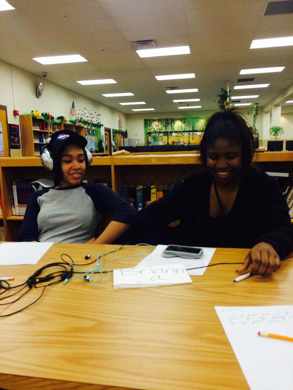 Two teens wearing headphones looking down at a smartphone on a piece of paper on a wooden table in a library