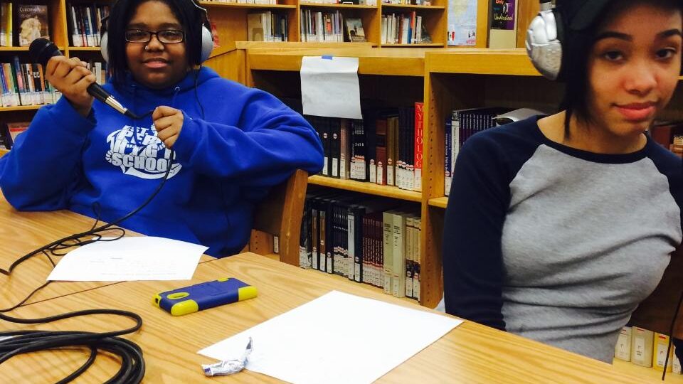 FullSizeRender (3) Two teens, one wearing headphones, looking down at a smartphone on a piece of paper on a table in a library