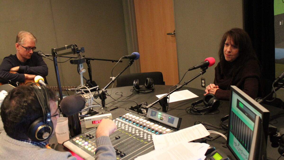 An adult sitting holding an acoustic guitar and an adult wearing headphones speaking into a microphone being interviewed by an adult wearing headphones sitting behind a microphone and a mixing board in a radio studio