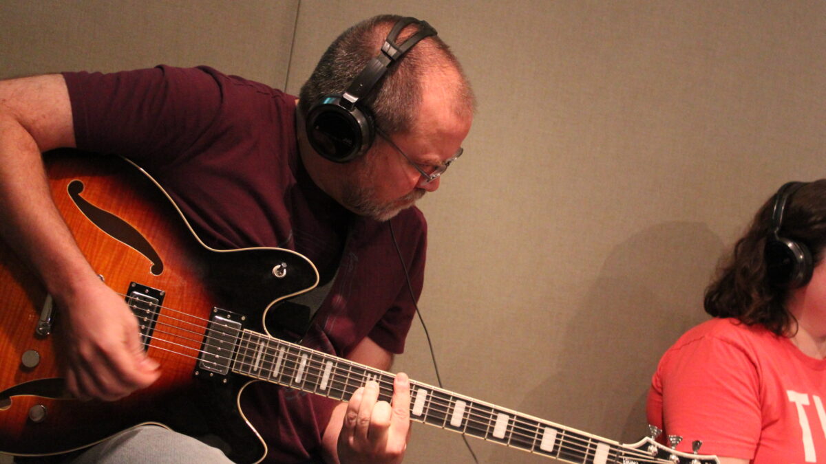 IMG_0636 An adult wearing headphones playing an acoustic guitar in a radio studio