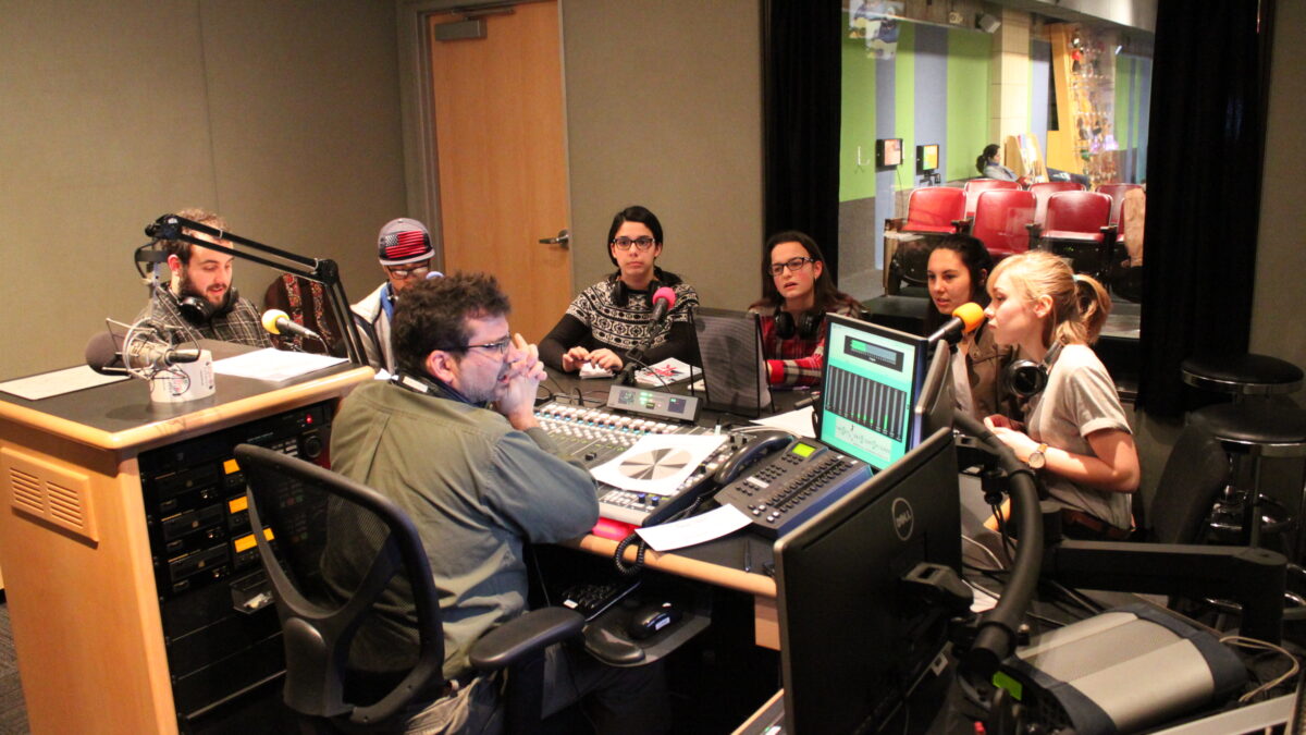 Six teens wearing headphones sitting behind microphones and an adult sitting behind a mixing board in a radio studio