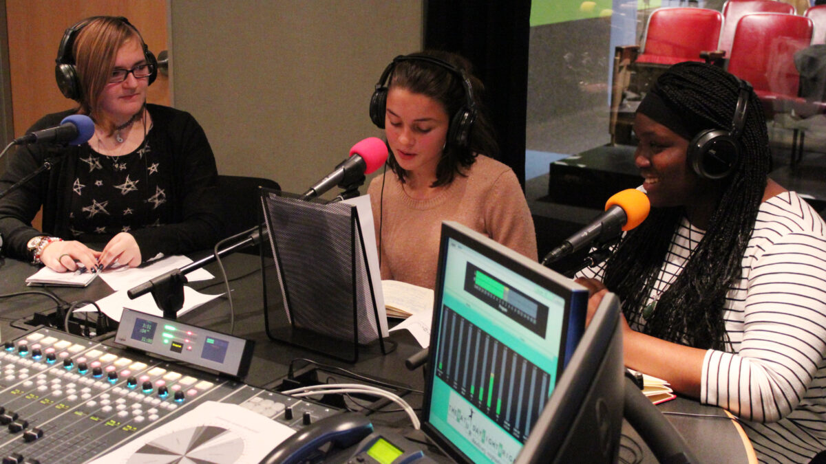 Three teens wearing headphones speaking into microphones in a radio studio