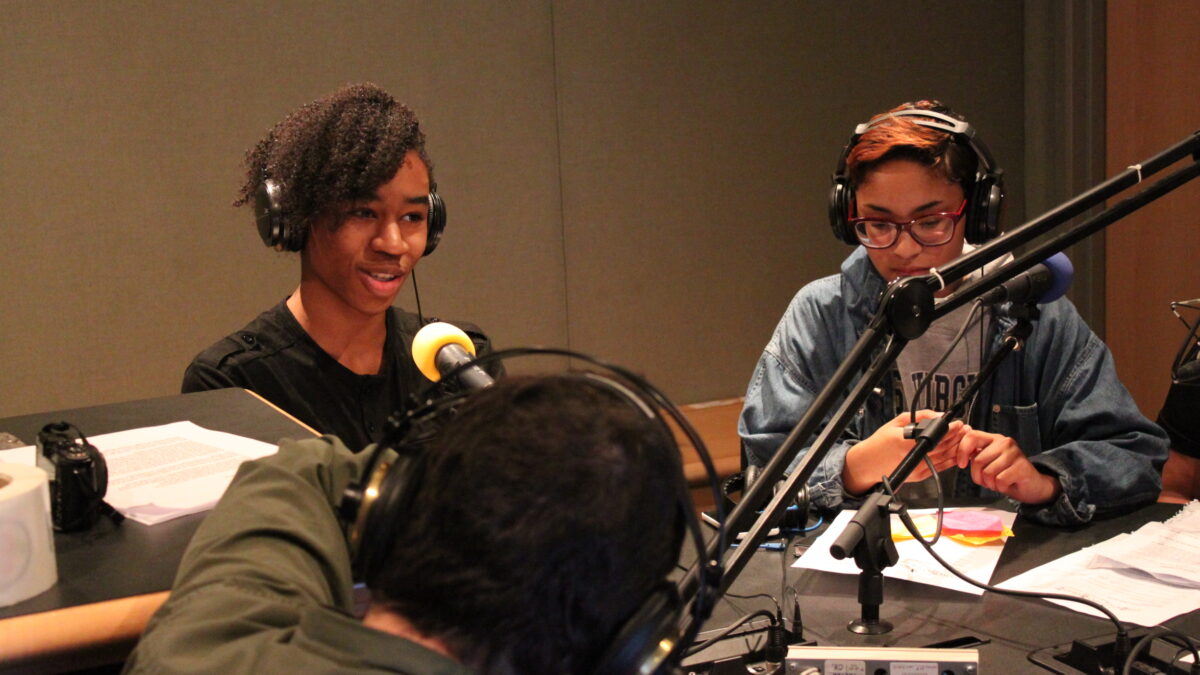 Two teens wearing headphones speaking into microphones and an adult wearing headphones sitting behind a mixing board in a radio studio