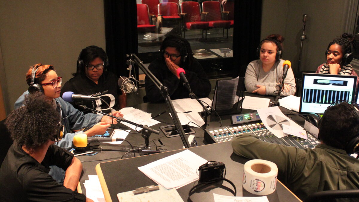 Six teens wearing headphones speaking into microphones and an adult wearing headphones sitting behind a mixing board in a radio studio