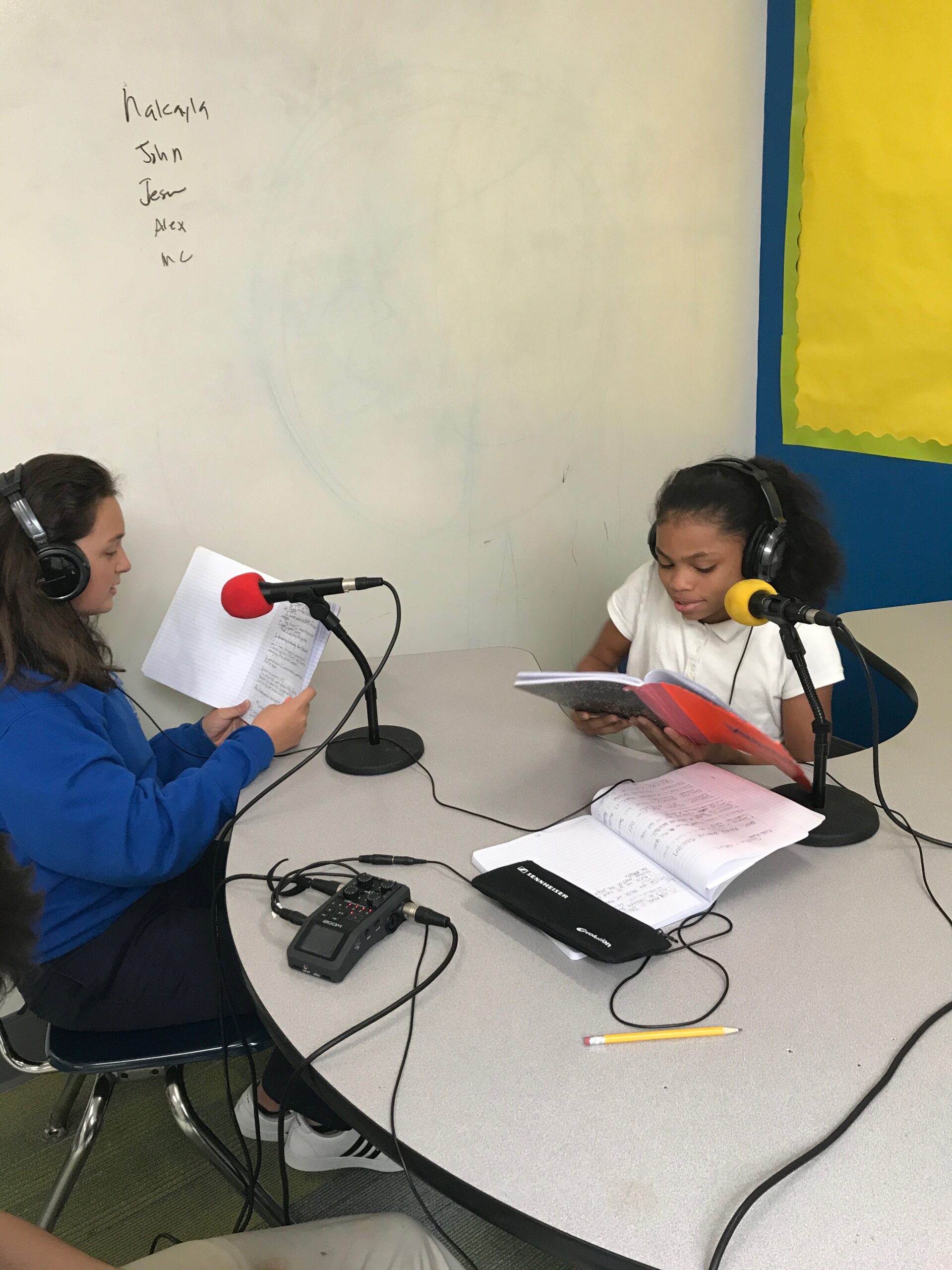 A youth looking at a notebook and an adult, both wearing headphones and speaking into microphones connected to a portable recorder on a table