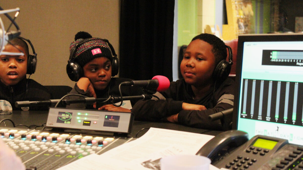 Three youth wearing headphones sitting behind microphones in a radio studio
