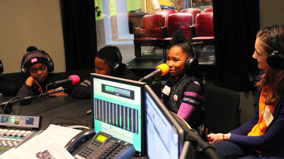 Three youth and an adult wearing headphones speaking into microphones in a radio studio