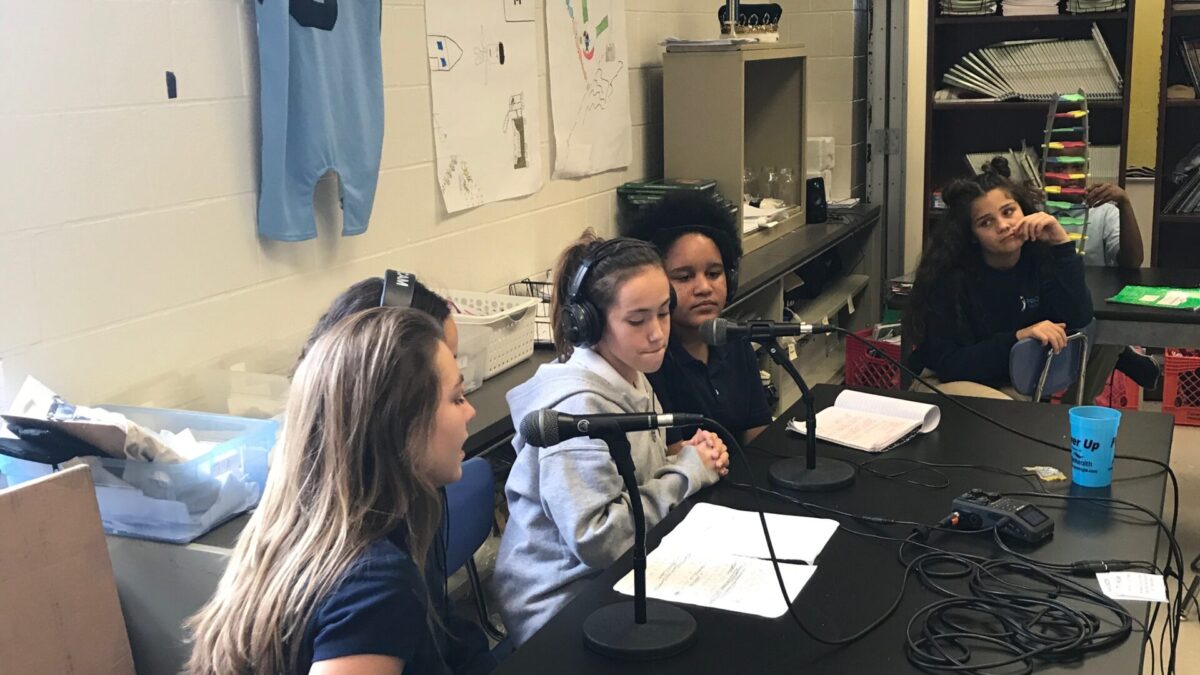 Five youth sitting around microphones on a long black table, three wearing microphones
