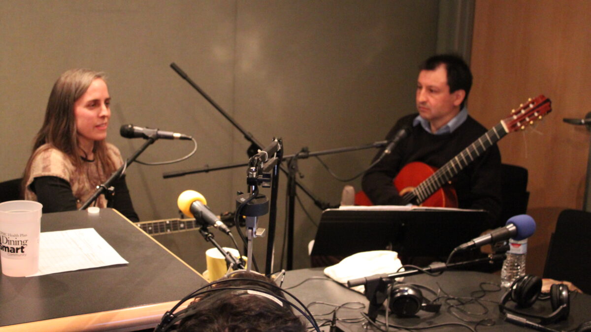 IMG_0834 An adult sitting behind a microphone holding a string instrument and an adult holding an acoustic guitar sitting behind a microphone in a radio studio