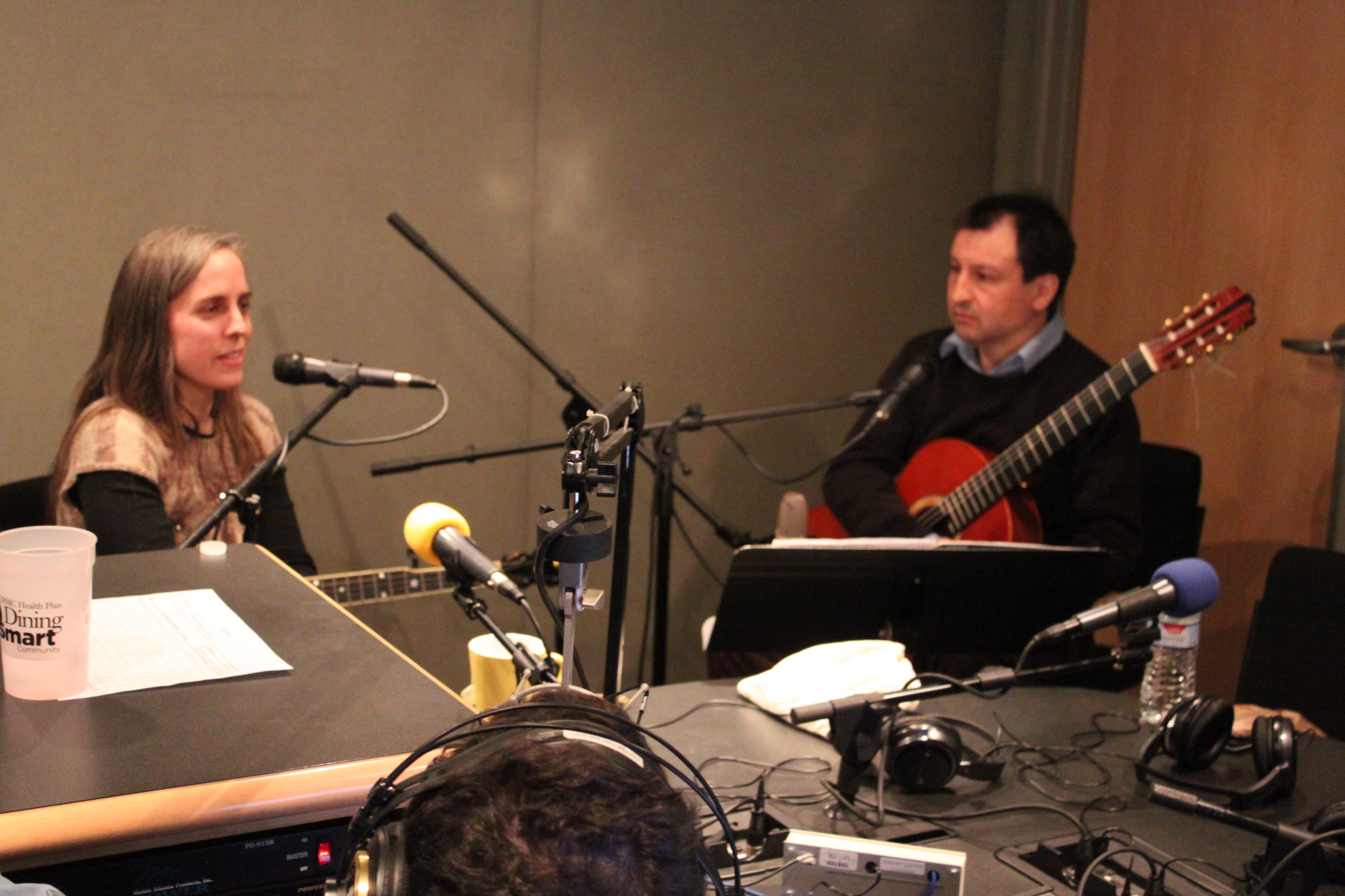 An adult sitting behind a microphone holding a string instrument and an adult holding an acoustic guitar sitting behind a microphone in a radio studio
