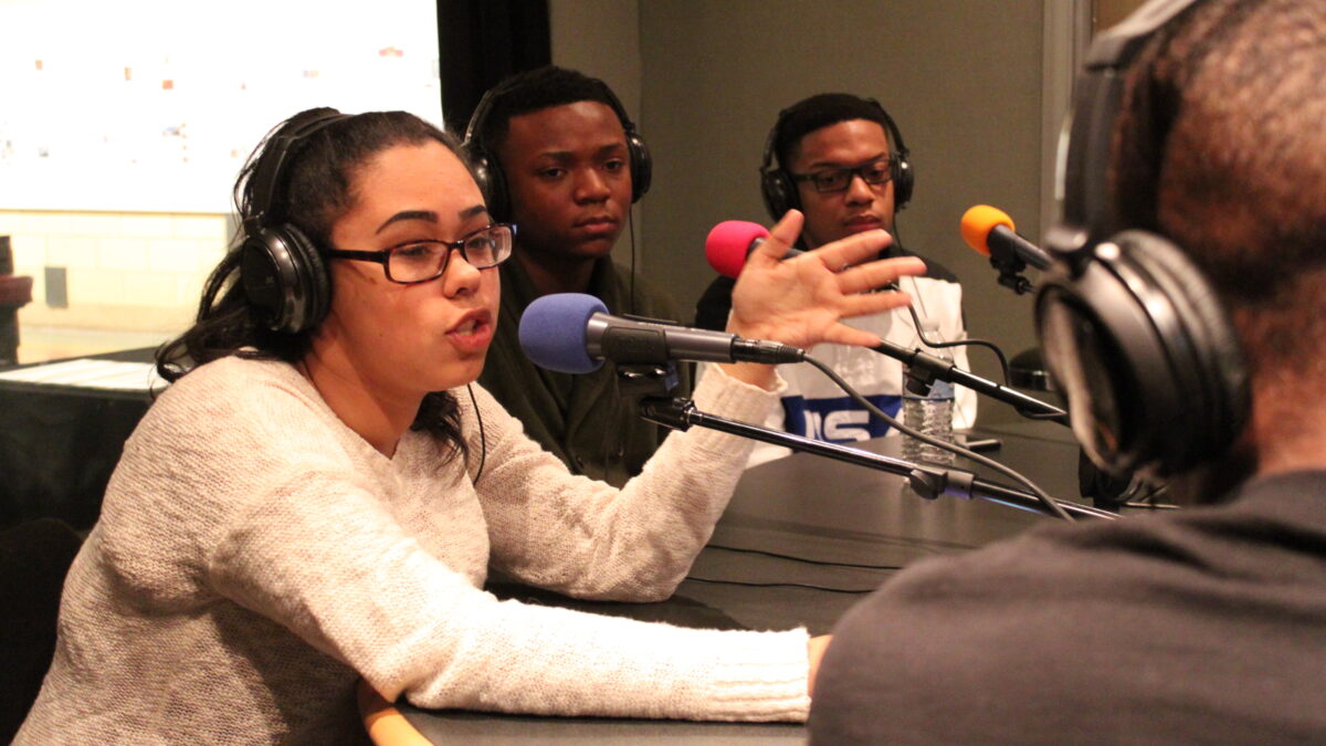 Four teens wearing headphones speaking into microphones in a radio studio