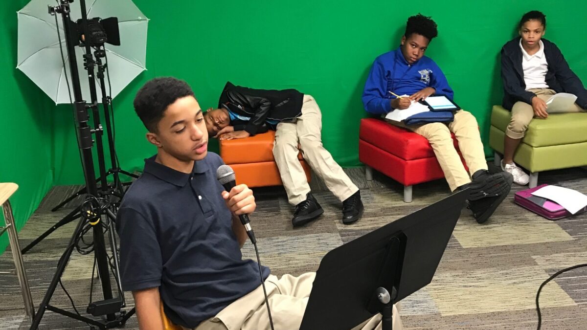 A youth speaking into a microphone looking at a music stand as three youth listen in the background