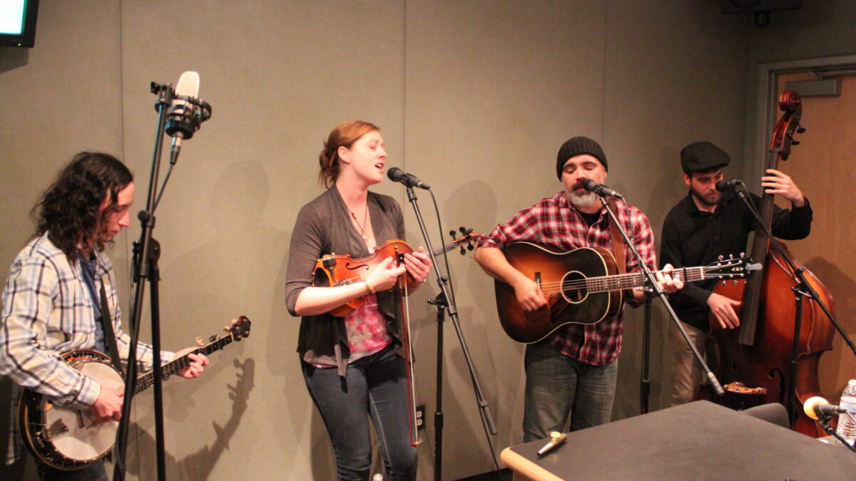 An adult playing banjo, an adult playing fiddle and singing into a microphone, an adult playing acoustic guitar, and an adult playing upright bass in a radio studio