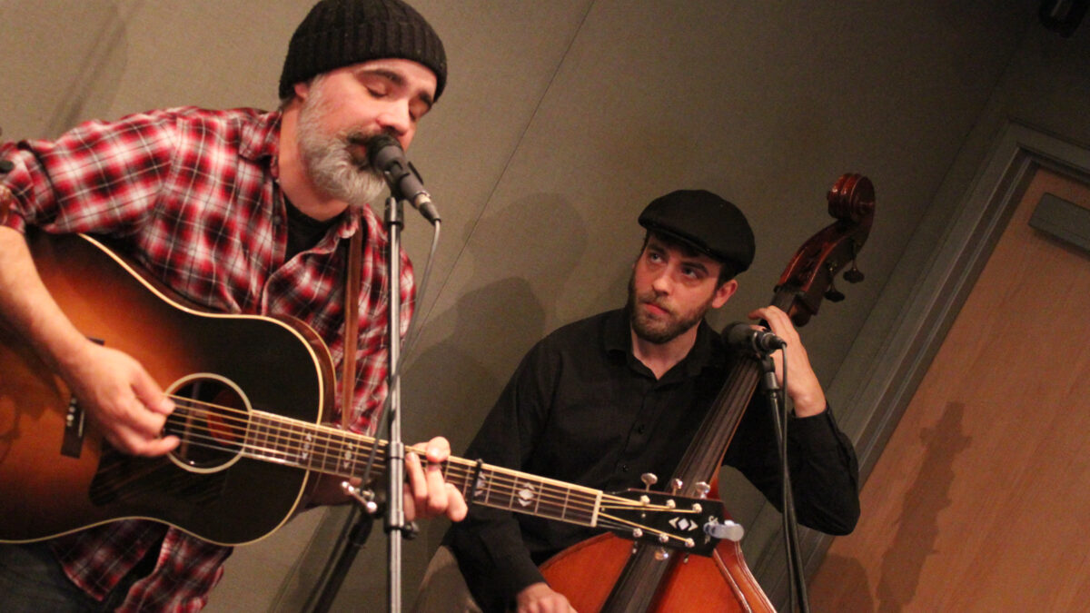 An adult playing acoustic guitar and singing into a microphone and an adult playing upright bass in a radio studio