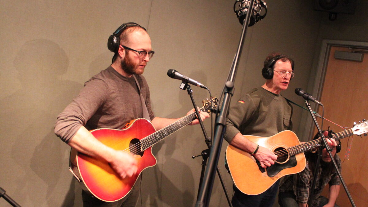 Two adults wearing headphones playing acoustic guitars in a radio studio