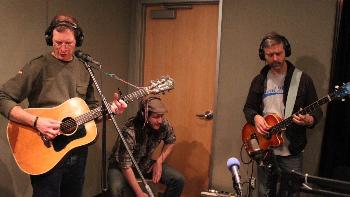 An adult wearing headphones playing acoustic guitar, an adult wearing headphones playing cajon, and an adult wearing headphones playing electric bass in a radio studio