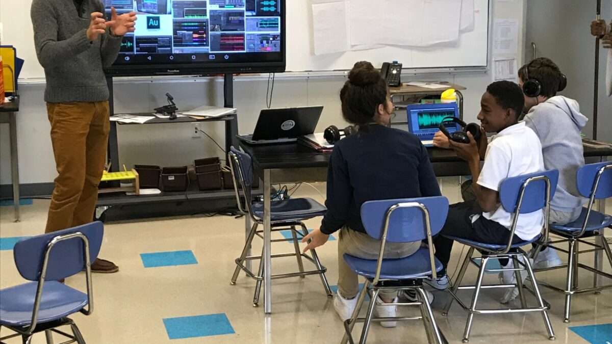 An adult presenting in front of a tv screen in a classroom as three youth sitting behind a laptop computer listen
