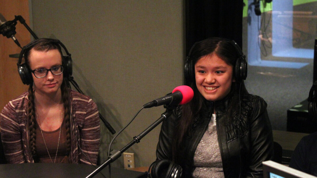 Two teens wearing headphones speaking into microphones in a radio studio