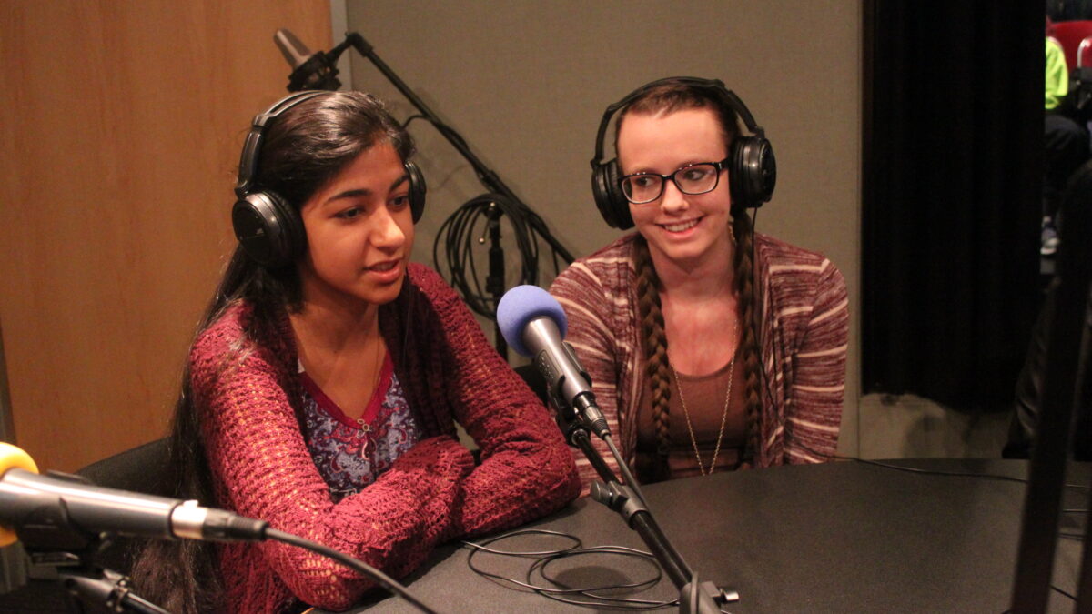 Two teens wearing headphones speaking into microphones in a radio studio