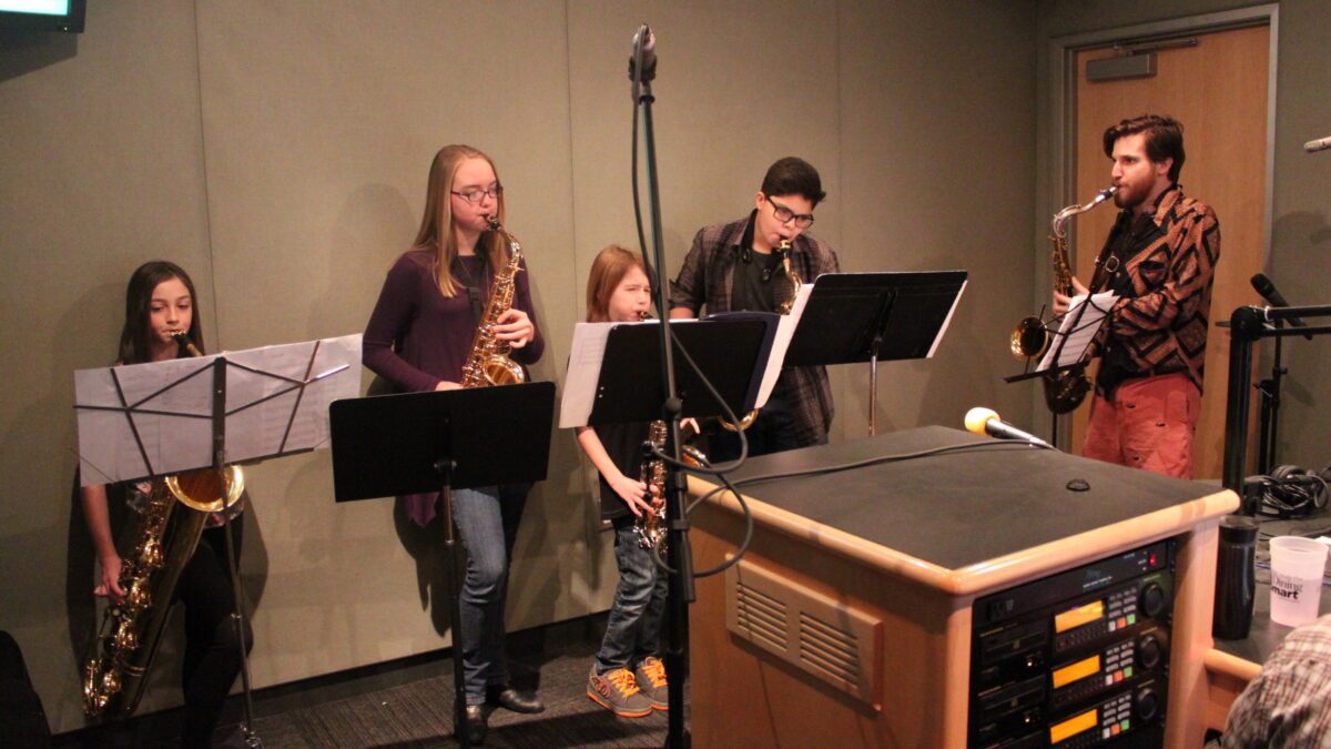 Two youth playing alto saxophone, one youth playing baritone saxophone, a youth playing tenor saxophone, and one adult playing tenor saxophone, all looking at sheet music on music stands in a radio studio