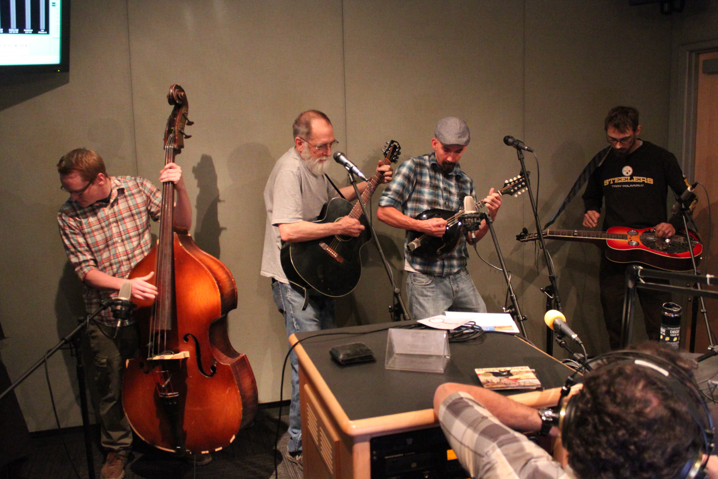 An adult playing an upright bass, an adult playing an acoustic guitar, an adult playing a mandolin, and an adult playing a dobro as another adult wearing headphones listens in a radio studio