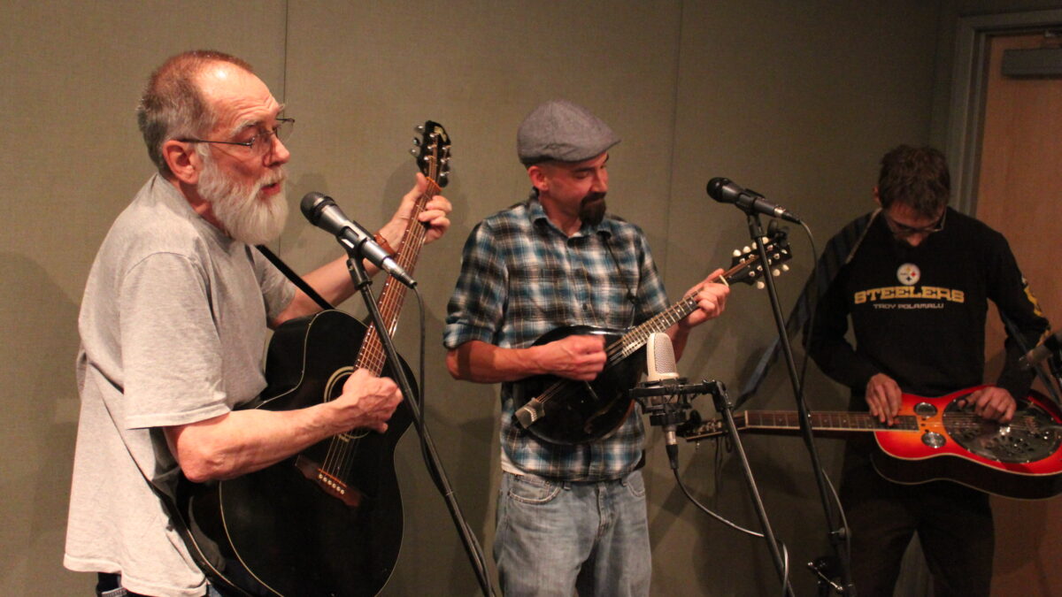 An adult playing acoustic guitar and singing into a microphone, an adult playing mandolin, and an adult playing dobro in a radio studio