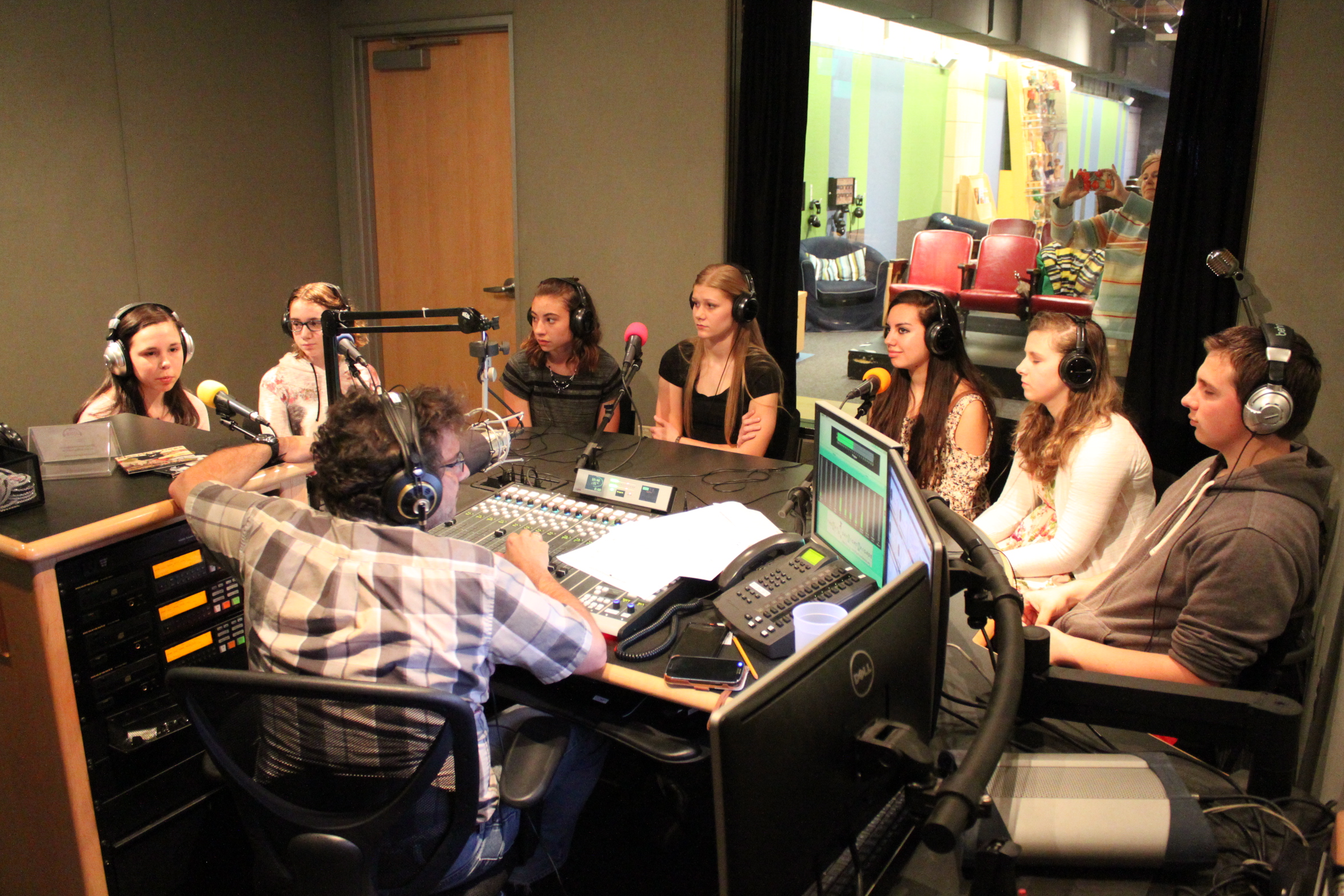 Seven youth wearing headphones speaking into microphones being interviewed by an adult wearing headphones sitting behind a microphone and a mixing board in a radio studio