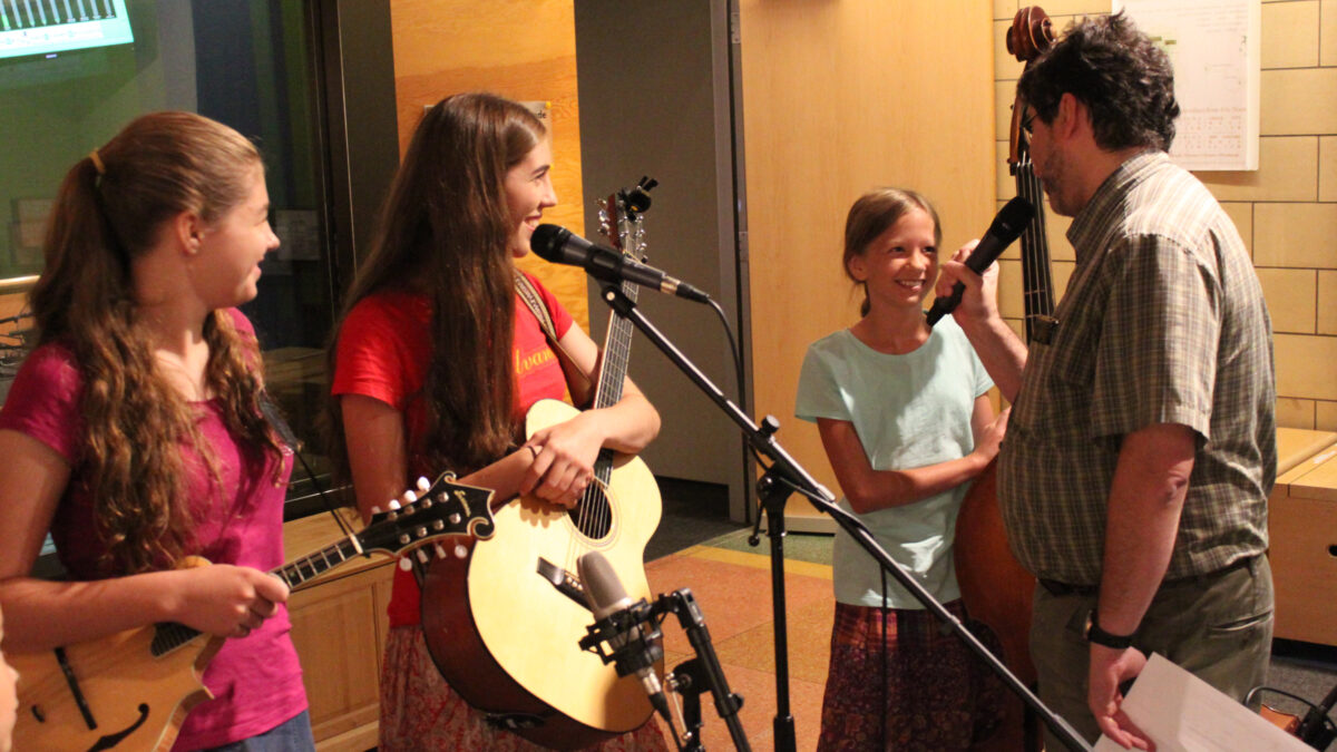 IMG_1459EV A youth holding a mandolin, a youth holding an acoustic guitar, and a youth holding an upright bass being interviewed by an adult holding and speaking into a microphone