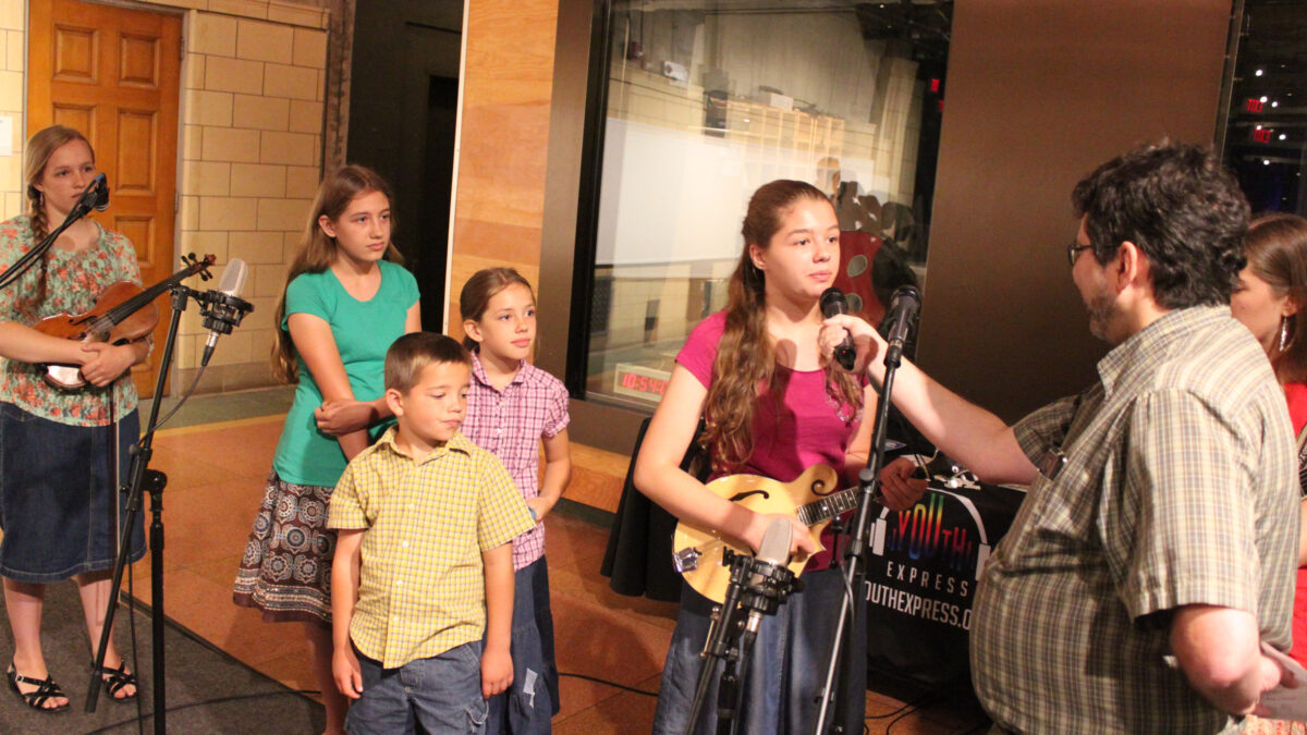 IMG_1460EV A youth holding a violin, three youth standing behind a microphone, and a youth holding a mandolin speaking into a microphone being held by an adult