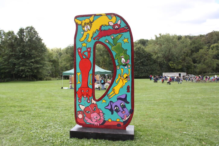 A large light blue art installation of the letter "D" with cartoon drawings on it in a large green field.