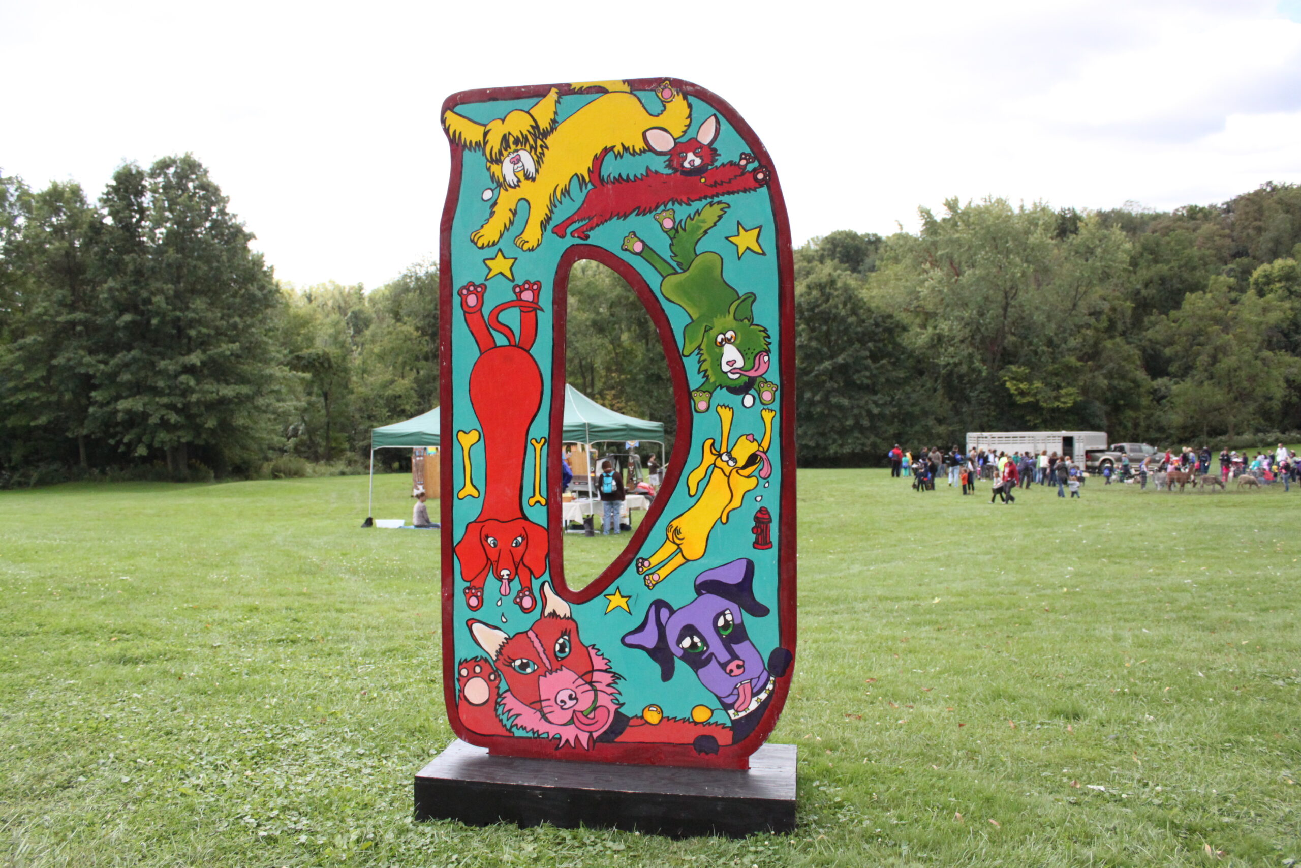 A large light blue art installation of the letter "D" with cartoon drawings on it in a large green field.