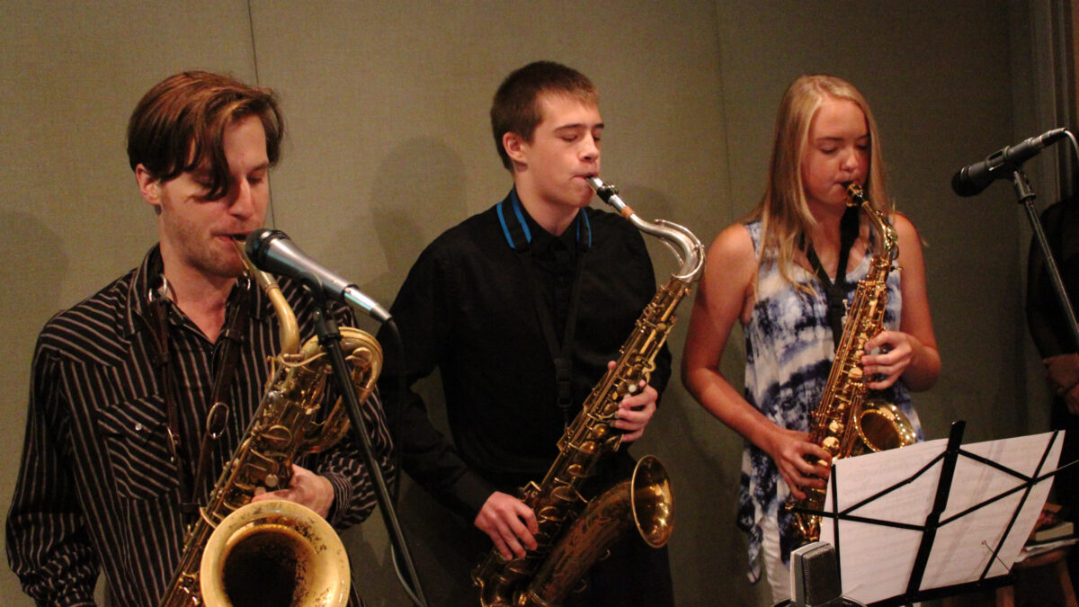 An adult playing a baritone saxophone, a teen playing a tenor saxophone, and a teen playing an alto saxophone, all looking at sheet music on a music stand in a radio studio